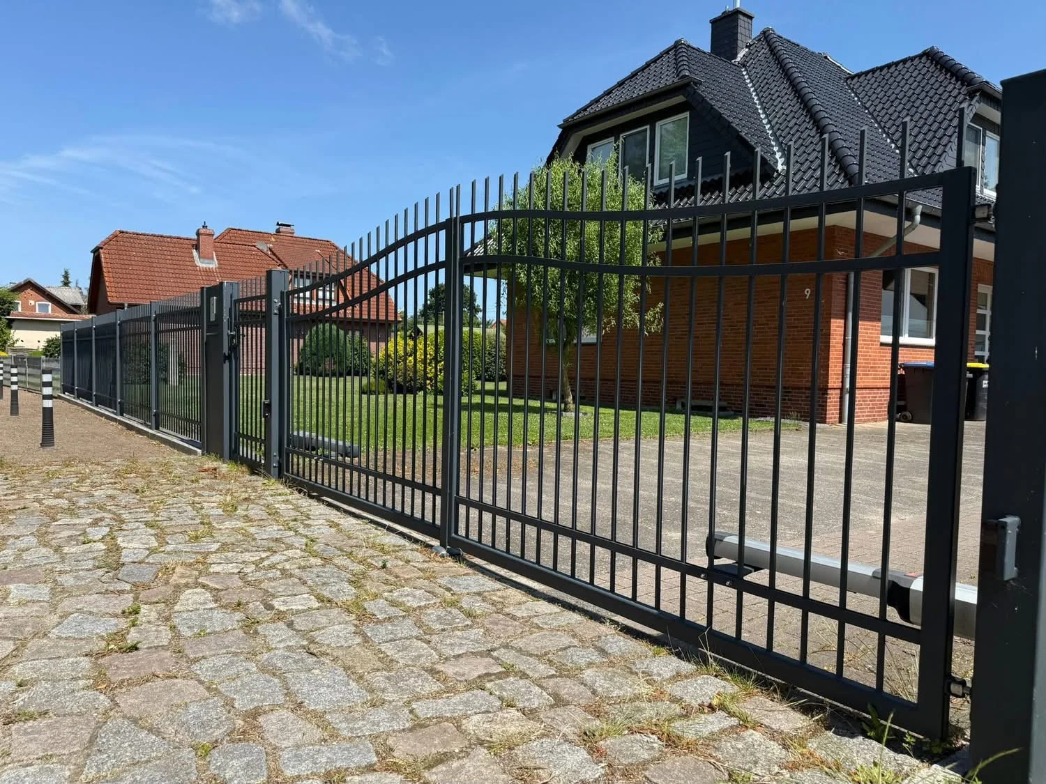 Zweiflügeliges Tor mit senkrechten Stäben und gebogener Oberkante an gepflasterter Einfahrt